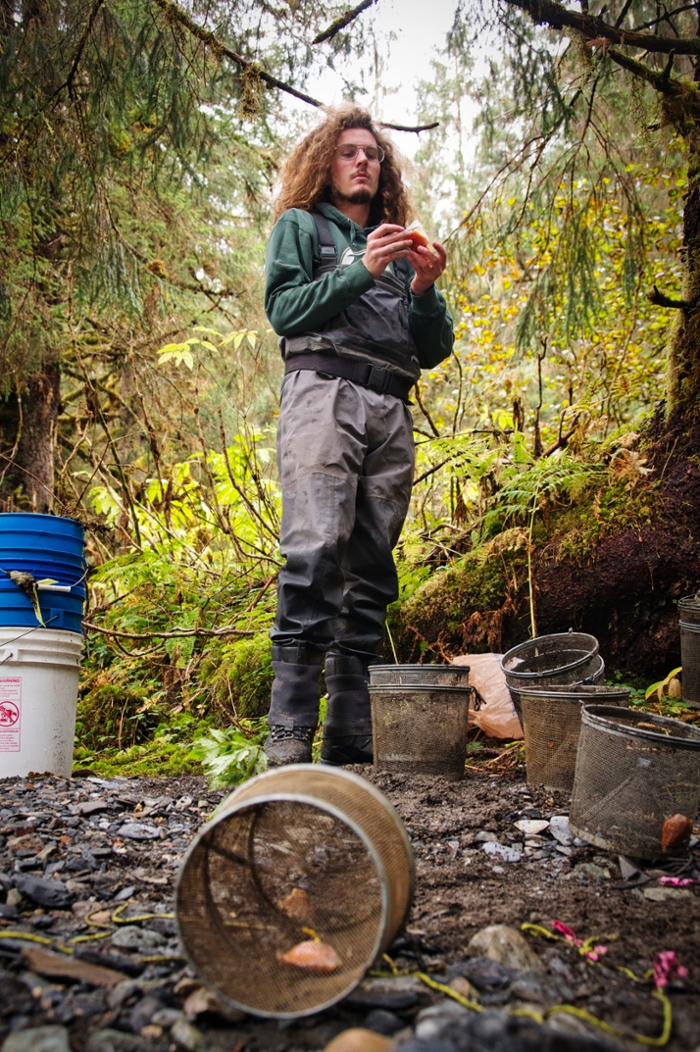 a man stands on a river bank in the forest holding a small container of fish eggs, with wire baskets at his feet