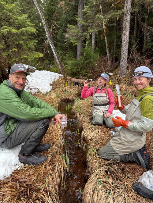 ACRC interns and director Jason hold stream sampling equipment while sitting by a creek
